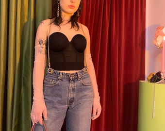 Könnte beinhalten: Eine Frau trägt ein schwarzes Korsett-Oberteil mit einer goldenen Kette, durchsichtige Handschuhe und eine blaue Jeans. Sie hält eine rosa und lilafarbene Tragetasche mit dem Prada-Logo.