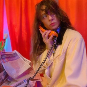 May include: A woman with long brown hair is talking on a vintage orange rotary phone. She is wearing a white blazer and a black choker necklace. The phone cord is coiled around her arm. She is sitting in front of a red curtain.