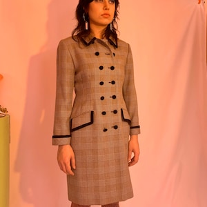 May include: A woman wearing a plaid double-breasted coat with black velvet trim. The coat has a fitted silhouette and a collar with a black velvet trim. The woman is wearing black and white patterned tights.