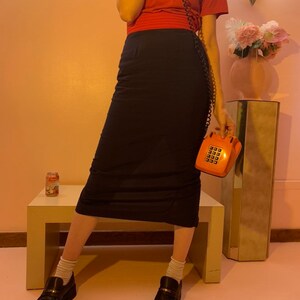 May include: A woman wearing a red t-shirt and a black skirt is holding a vintage orange rotary phone. She is standing on a wooden bench with a can of soda on the surface. The background is a pink wall.