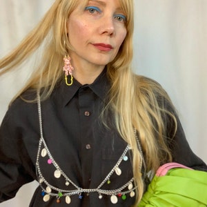 May include: A woman with long blonde hair wearing a black shirt with a silver chain bra top with colorful beads and a green purse.