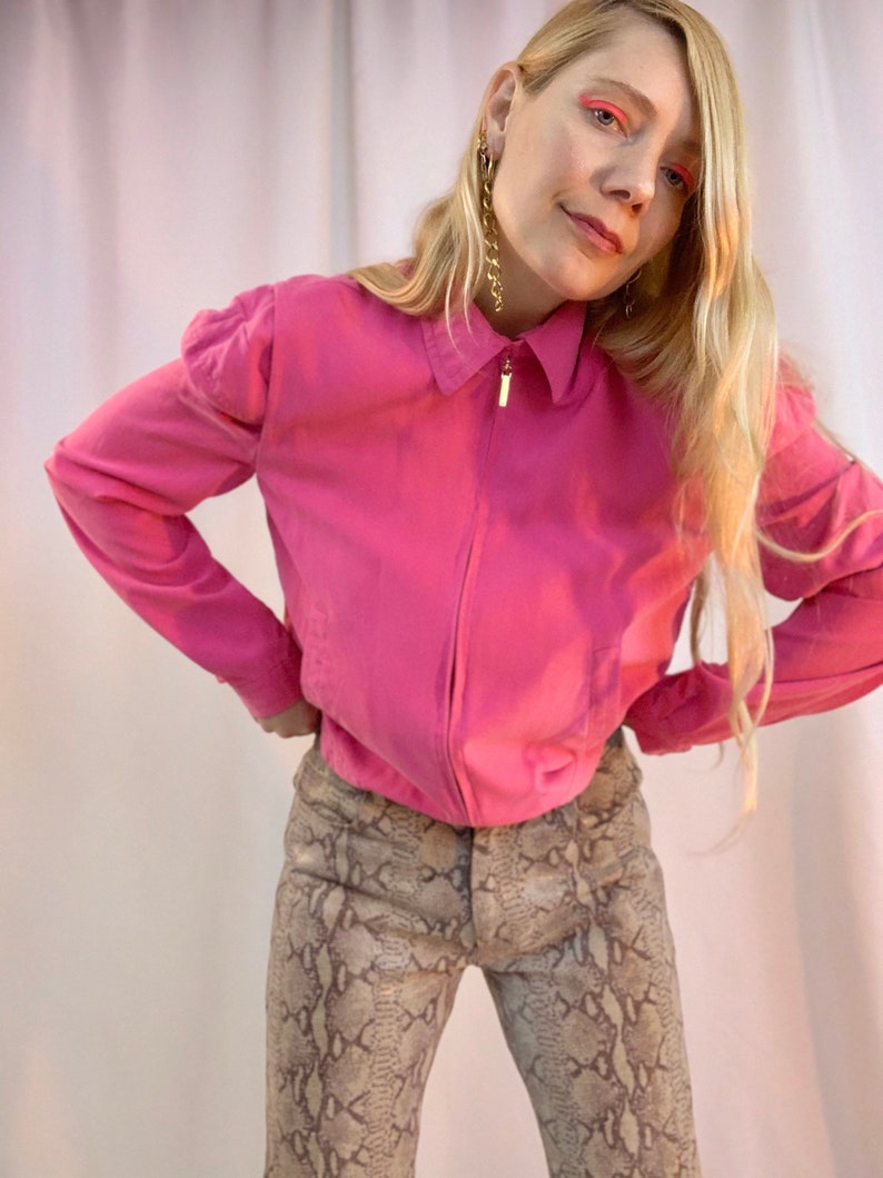May include: A woman wearing a pink collared shirt with a zipper and a pair of snake print pants.
