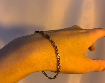 Puede incluir: Un brazalete de cadena dorada con un diseño trenzado.