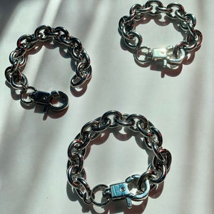 May include: Three silver chain bracelets with a clasp closure. The bracelets are made of thick, chunky links.