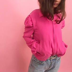 May include: A hot pink bomber jacket with puffed sleeves and a zipper closure. The jacket is worn over a pair of light blue jeans.