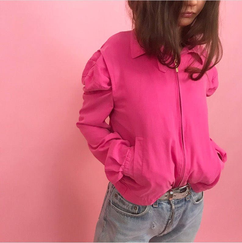 May include: A hot pink bomber jacket with puffed sleeves and a zipper closure. The jacket is worn over a pair of light blue jeans.