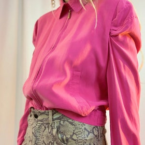 May include: A pink, long-sleeved shirt with a zipper closure and puffed sleeves. The shirt is worn over a pair of brown and white snake-print pants.