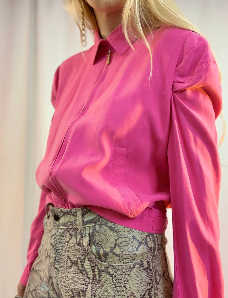 May include: A pink, long-sleeved shirt with a zipper closure and puffed sleeves. The shirt is worn over a pair of brown and white snake-print pants.