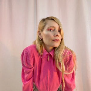 May include: A woman with long blonde hair is wearing a pink collared shirt with a zipper. She is wearing gold chain earrings and has pink eyeshadow.