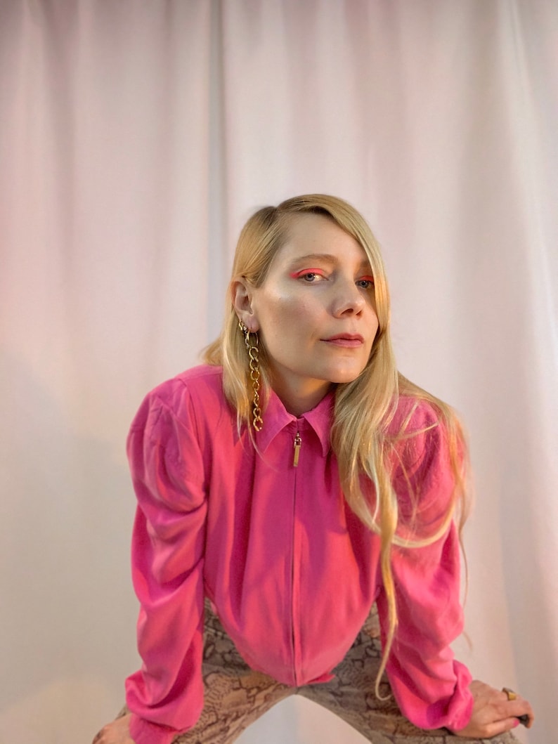 May include: A woman with long blonde hair is wearing a pink collared shirt with a zipper. She is wearing gold chain earrings and has pink eyeshadow.