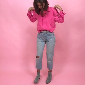 May include: A woman wearing a bright pink long-sleeved shirt with a collared neckline, light blue jeans with distressed details, and gray ankle boots.