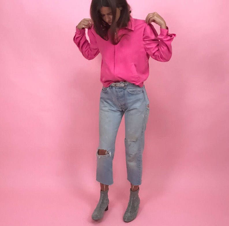 May include: A woman wearing a bright pink long-sleeved shirt with a collared neckline, light blue jeans with distressed details, and gray ankle boots.
