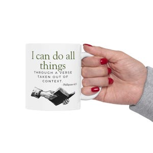 Peut inclure: Mug en céramique blanche avec l'inscription "I can do all things" en vert, ainsi qu'une citation de Philippiens 4:13. La tasse présente également une illustration en noir et blanc d'une main tenant un livre.