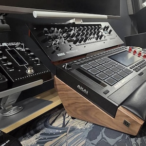 Może przedstawiać: Konfiguracja studia z wyposażeniem do produkcji muzyki elektronicznej. Obraz przedstawia czarny Akai MPC One, Korg Minilogue XD i inne interfejsy audio. Sprzęt jest zamontowany na drewnianym stojaku, z czarną skórzaną podpórką pod nadgarstek.
