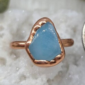 May include: A copper ring with a light blue aquamarine stone set in the centre. The ring is sitting on a white surface.