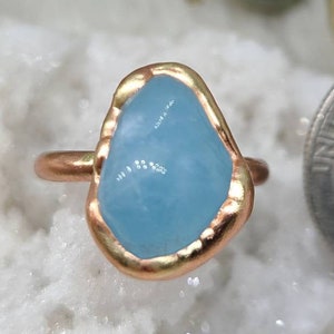 Op de afbeelding: Een ring met een lichtblauwe aquamarijn-steen gezet in een gouden metalen band. De steen heeft een onregelmatige vorm en een ruwe, natuurlijke uitstraling.