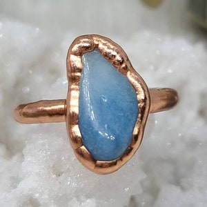Puede incluir: Un anillo de cobre con una piedra de aguamarina azul claro engastada en el centro. La piedra tiene una forma irregular y un aspecto &aacute;spero y natural.