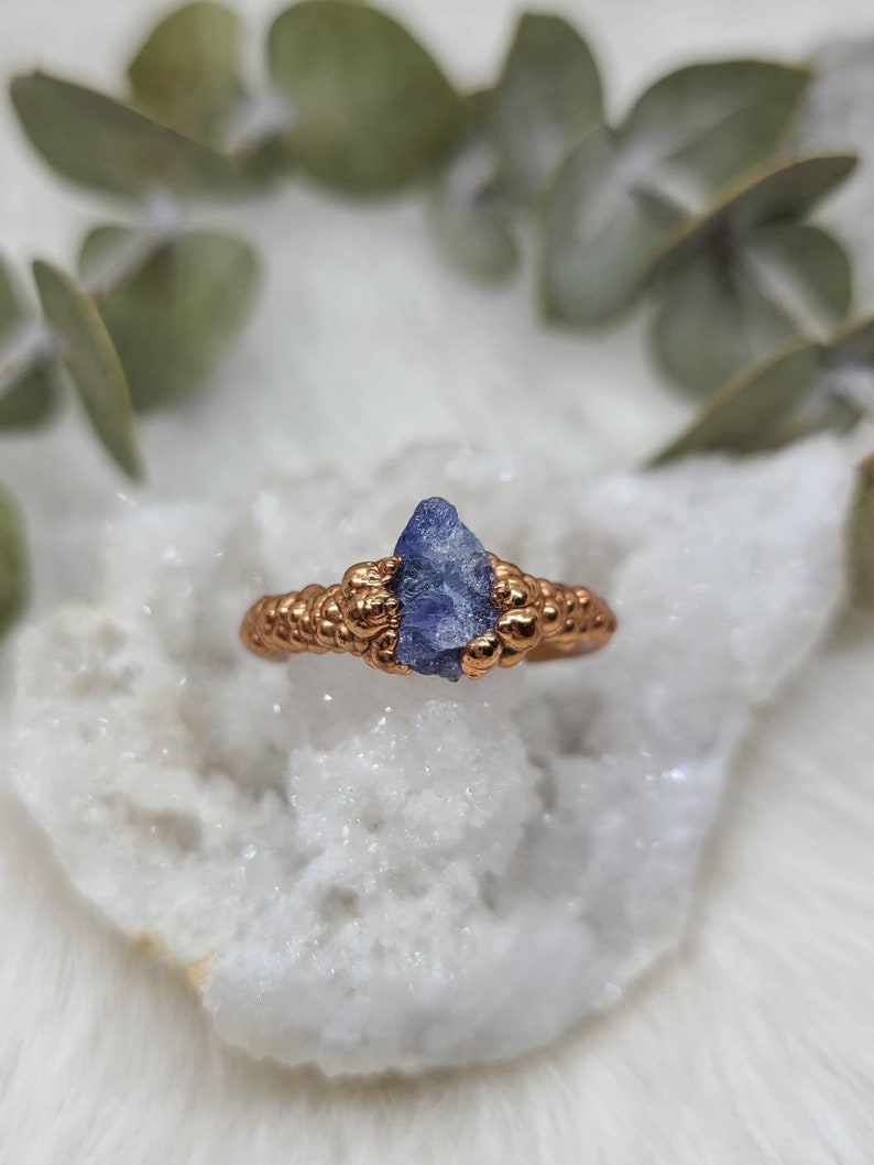 Puede incluir: Un anillo de oro con una piedra preciosa azul engastada en el centro. El anillo est&aacute; hecho de alambre que se envuelve alrededor de la piedra y luego se retuerce para crear una banda texturizada.