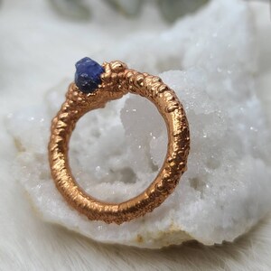 Puede incluir: Un anillo de cobre con una superficie texturizada y una piedra preciosa azul engastada en la banda.