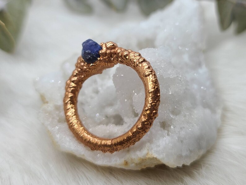 Puede incluir: Un anillo de cobre con una superficie texturizada y una piedra preciosa azul engastada en la banda.