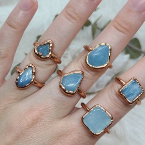 Puede incluir: Cinco anillos de cobre con piedras de aguamarina de diferentes formas. Los anillos est&aacute;n en una mano.