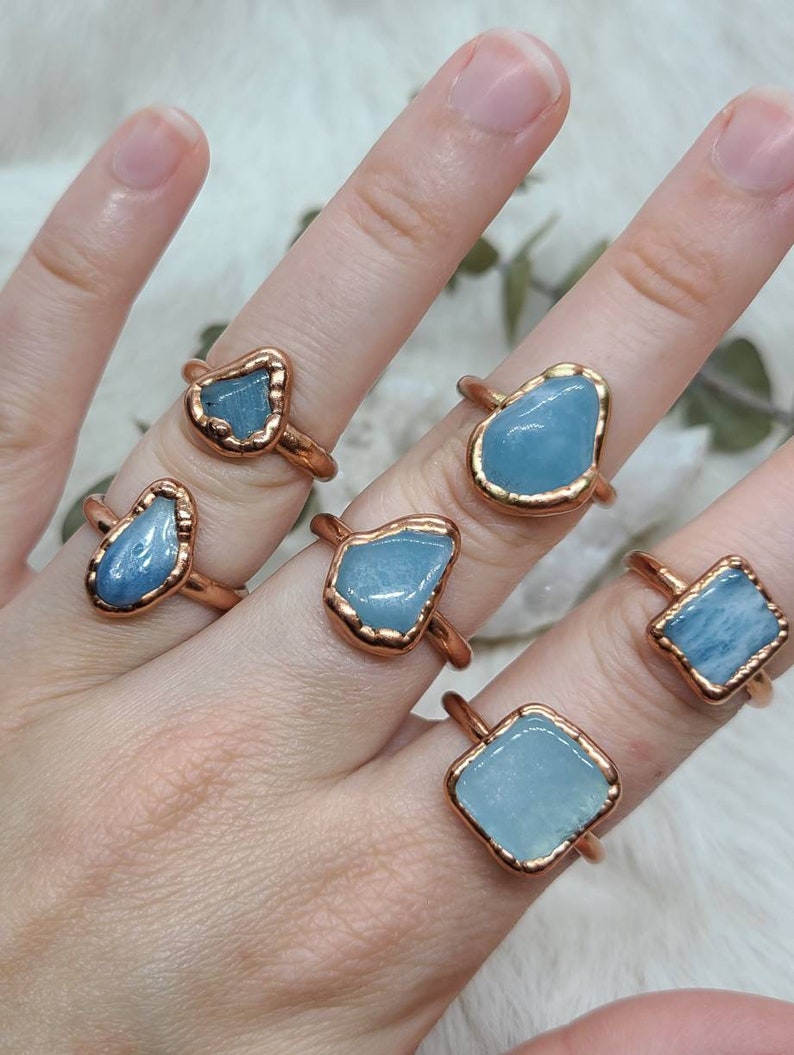 Puede incluir: Cinco anillos de cobre con piedras de aguamarina de diferentes formas. Los anillos est&aacute;n en una mano.
