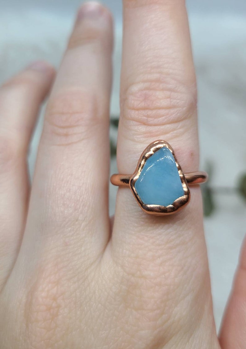 May include: A copper ring with a light blue aquamarine stone set in the centre. The ring is on a finger.