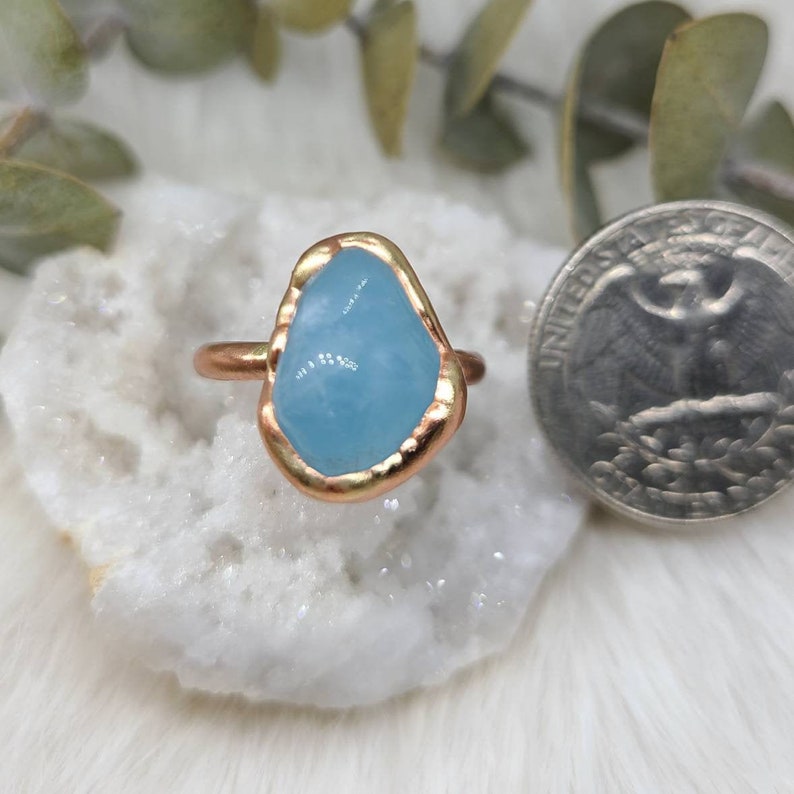 Op de afbeelding: Een ring met een lichtblauwe aquamarijn-steen gezet in een gouden metalen band. De steen heeft een onregelmatige vorm en een ruwe, natuurlijke uitstraling.