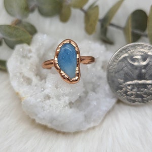 Puede incluir: Un anillo de cobre con una piedra preciosa azul claro engastada en el centro. El anillo est&aacute; sobre un fondo blanco.