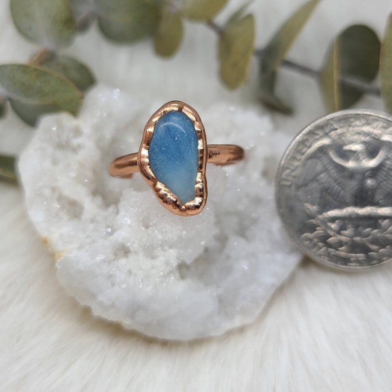 Puede incluir: Un anillo de cobre con una piedra preciosa azul claro engastada en el centro. El anillo est&aacute; sobre un fondo blanco.