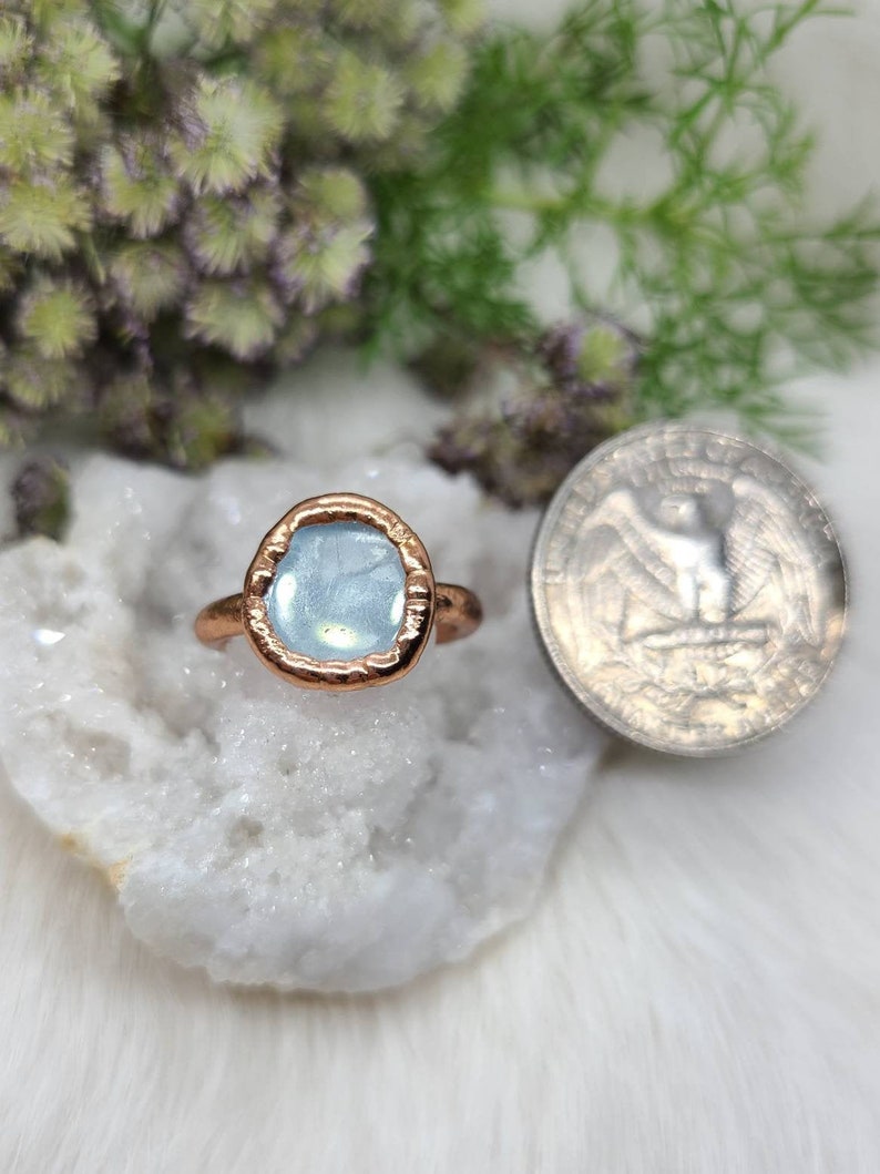 Puede incluir: Un anillo de cobre con una piedra preciosa azul claro engastada en un engaste de bisel. El anillo se muestra sobre un fondo blanco.