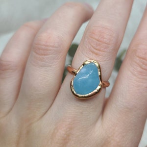 Op de afbeelding: Een gouden ring met een lichtblauwe edelsteen gezet in een gehamerde gouden zetting. De ring wordt gedragen aan een vinger.
