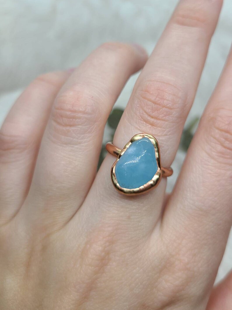 Op de afbeelding: Een gouden ring met een lichtblauwe edelsteen gezet in een gehamerde gouden zetting. De ring wordt gedragen aan een vinger.