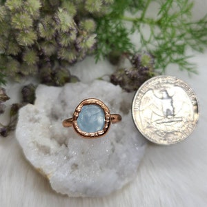 Puede incluir: Un anillo de cobre con una piedra de aguamarina azul claro engastada en un bisel de cobre martillado. El anillo est&aacute; sobre un cristal blanco.
