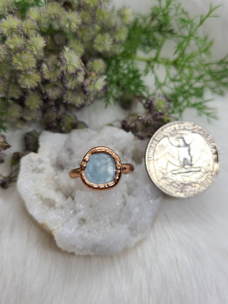 Puede incluir: Un anillo de cobre con una piedra de aguamarina azul claro engastada en un bisel de cobre martillado. El anillo est&aacute; sobre un cristal blanco.