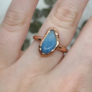 Puede incluir: Un anillo de cobre con una piedra preciosa azul engastada en un engaste de bisel. El anillo est&aacute; en un dedo.