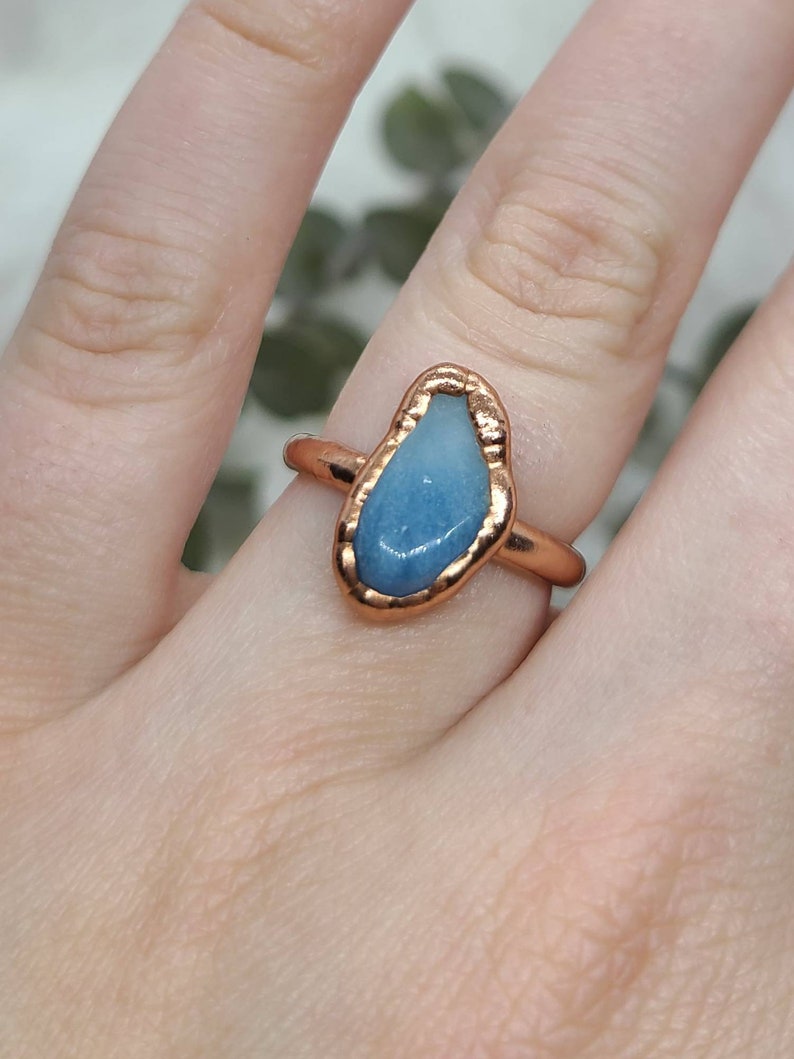 Puede incluir: Un anillo de cobre con una piedra preciosa azul engastada en un engaste de bisel. El anillo est&aacute; en un dedo.