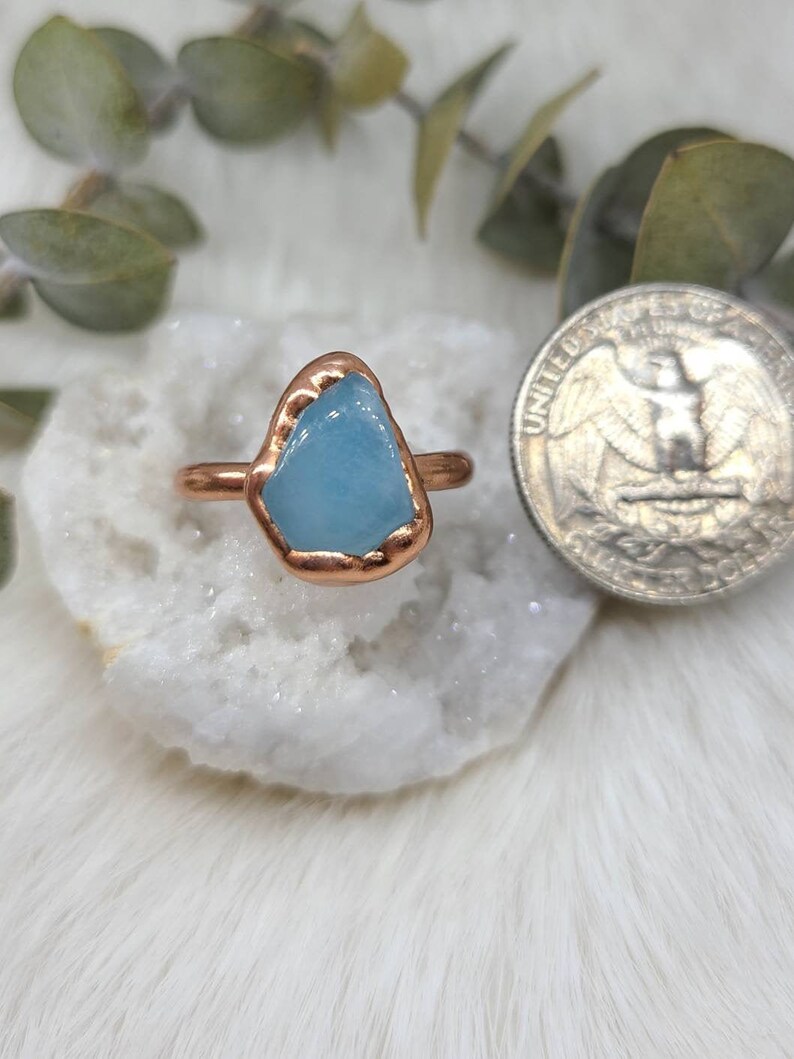 May include: A copper ring with a light blue aquamarine stone set in the centre. The ring is sitting on a white surface.