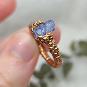 Puede incluir: Un anillo de cobre con una banda texturizada y una piedra de zafiro azul en bruto engastada en el centro.
