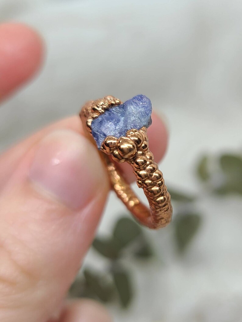 Puede incluir: Un anillo de cobre con una banda texturizada y una piedra de zafiro azul en bruto engastada en el centro.