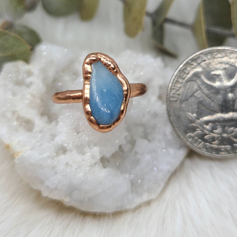 Puede incluir: Un anillo de cobre con una piedra de aguamarina azul claro engastada en el centro. La piedra tiene una forma irregular y un aspecto &aacute;spero y natural.