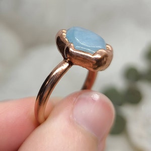 May include: A copper ring with a light blue gemstone set in a bezel setting. The ring is on a finger.