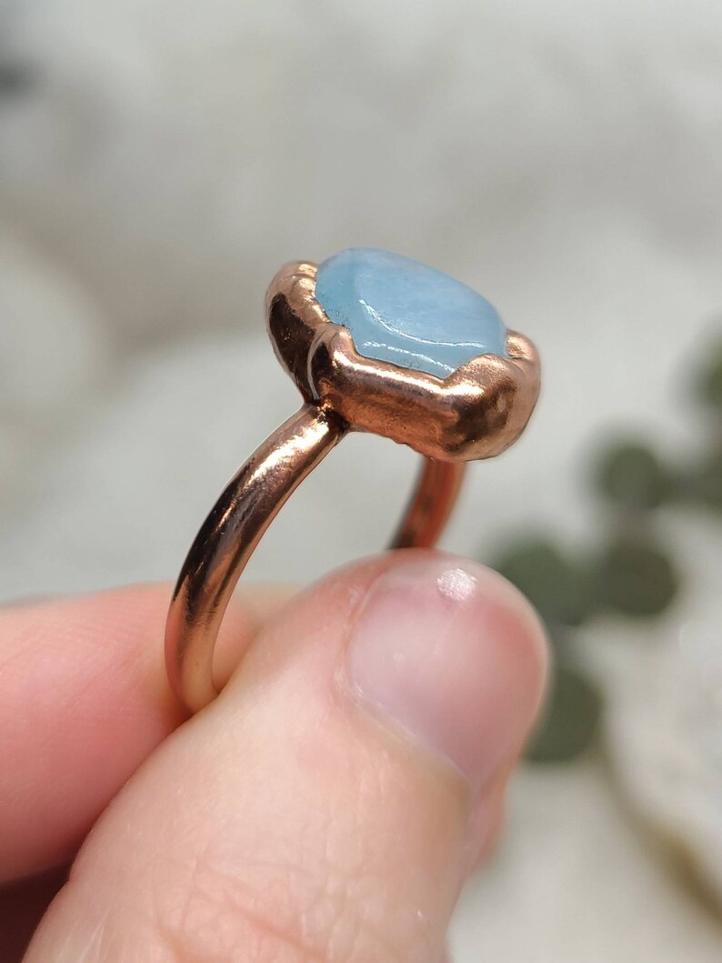 May include: A copper ring with a light blue gemstone set in a bezel setting. The ring is on a finger.