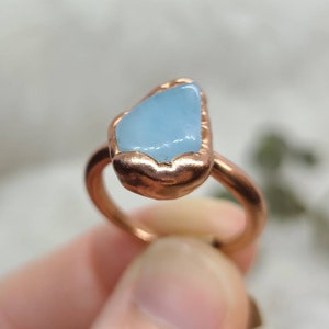 May include: A copper ring featuring a light blue aquamarine stone set in the centre. The ring is handmade and has a rustic, hammered finish.
