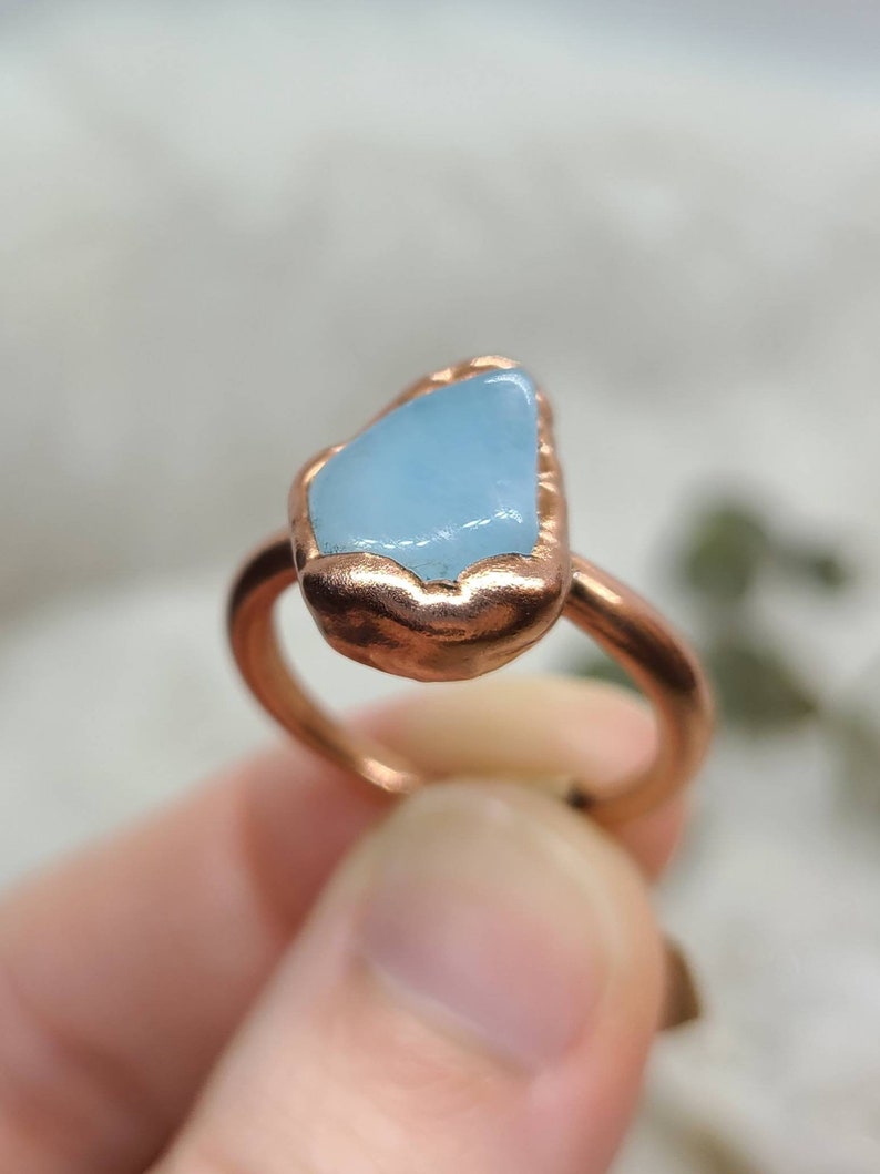 May include: A copper ring featuring a light blue aquamarine stone set in the centre. The ring is handmade and has a rustic, hammered finish.