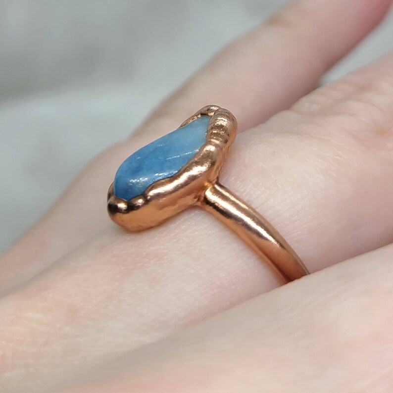Puede incluir: Un anillo de cobre con una piedra preciosa azul claro engastada en un engaste texturizado. El anillo se lleva en un dedo.