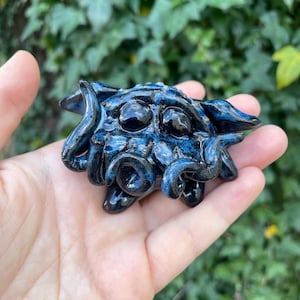 May include: A small, hand-painted ceramic sculpture of a blue and black creature with large eyes and tentacles. The creature has a speckled, textured surface.