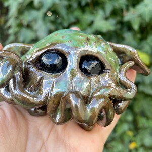 May include: A ceramic sculpture of a green and brown octopus with large black eyes. The sculpture has a textured surface and a glossy finish.