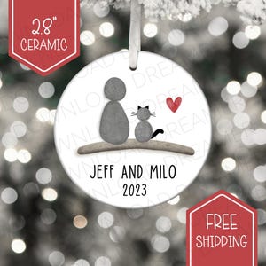 May include: A ceramic Christmas ornament, approximately 7.1 cm in diameter. It features a gray illustration of a person and a cat, with a red heart. The names "JEFF AND MILO" and the year "2023" are printed below the illustration.