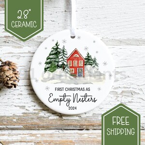 Peut inclure: Ornement de Noël en céramique de 7,1 cm. L'ornement rond blanc présente une maison rouge avec de la neige et des arbres à feuilles persistantes. Le texte indique "First Christmas as Empty Nesters 2024". Un ruban blanc est attaché pour l'accrocher.